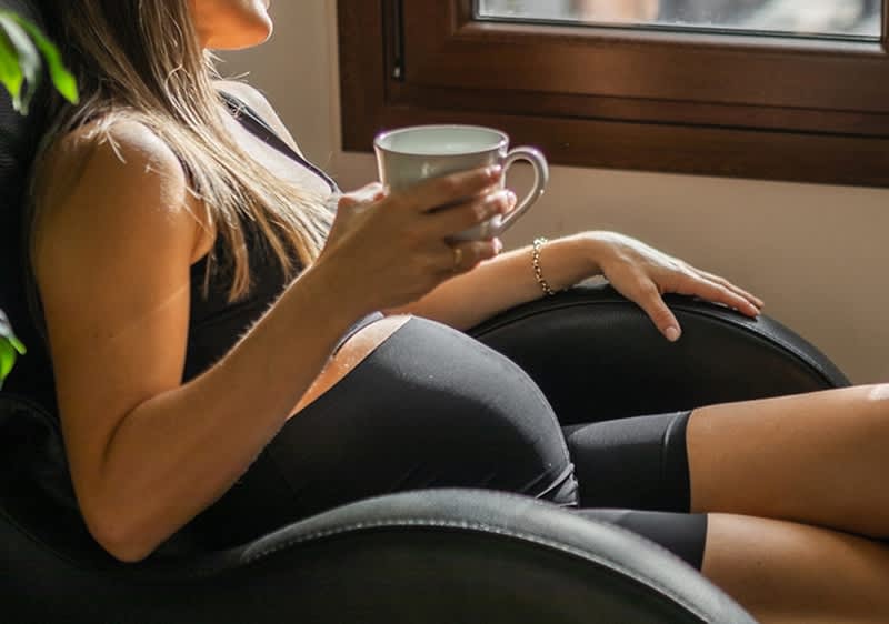 Schwangere Frau sitzt mit einer Tasse Tee in der Hand am Fenster
