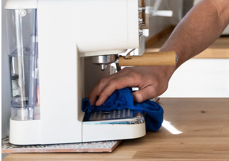 Eine Hand reinigt mit einem blauen Tuch die Tropfschale einer weißen Kaffeemaschine auf einem Holztisch