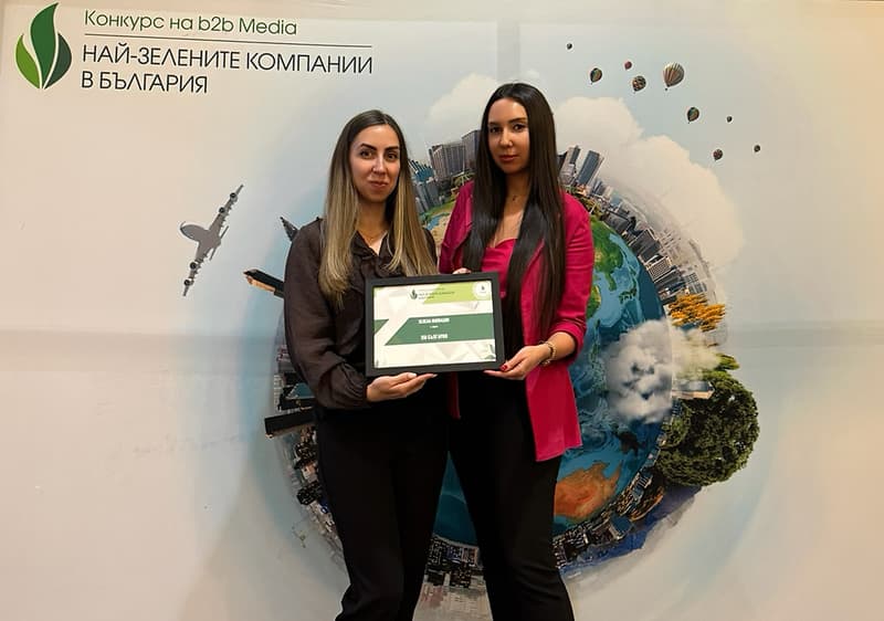 Две жени държат рамкирана награда пред плакат с планета и градски елементи