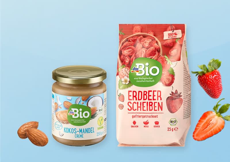 Links ein Glas dmBio Kokos-Mandel-Creme mit goldfarbenem Deckel und sichtbarem Etikett, rechts eine Packung dmBio Erdbeer Scheiben gefriergetrocknet 25 g mit rotem Design und abgebildeten Erdbeeren, Mandeln und aufgeschnittene Erdbeere