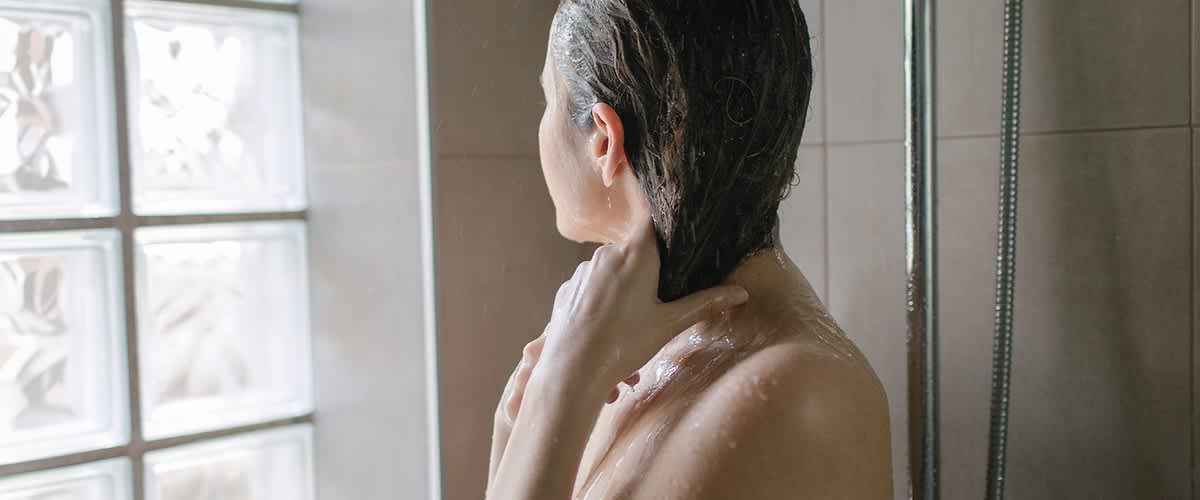 Eine Person steht unter der Dusche, mit nassen Haaren die Hände im Nacken und Wasser läuft über Schultern und Rücken