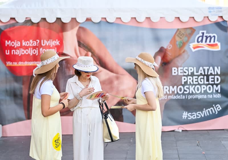 Alt-tekst: Promotori u žutim haljinama ispred DM štanda za besplatan pregled kože