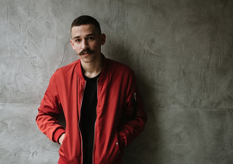 Ein Mann mit Schnurrbart und roter Jacke und vor grauer Wand