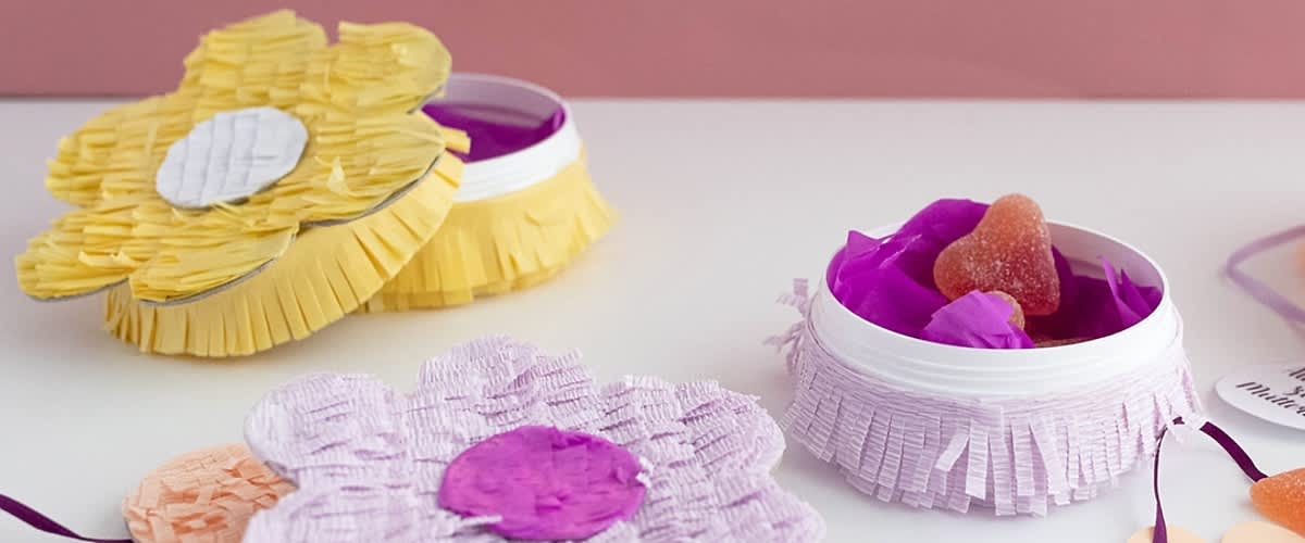 Rundes weißes Schälchen mit rosa Fransenband, gefüllt mit violetten Papierblüten und einem zuckerbestreuten Herzbonbon