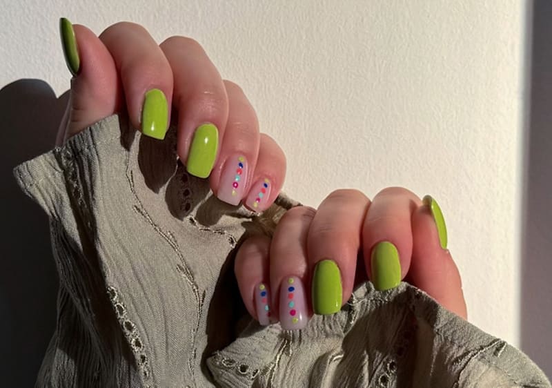 Zwei Hände mit hellgrün lackierten langen Nägeln, jeweils Ring- und Mittelfinger mit hellem Nageldesign und bunten Punkten, Hände greifen an einen hellgrünen Spitzenärmel
