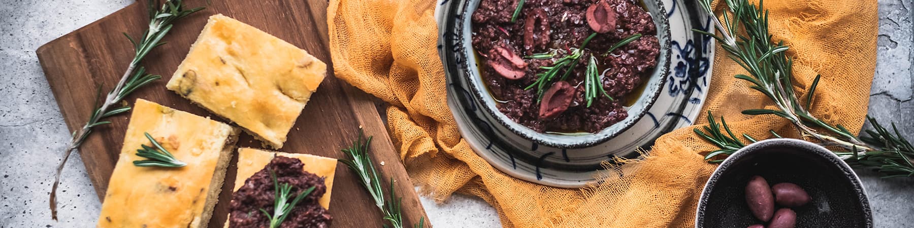 Focaccia s olivovou tapenádou, rozmarýnem a česnekem na dřevěném prkénku a talíři.
