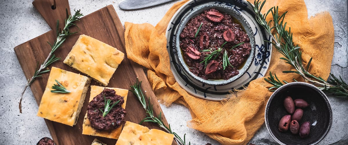 Focaccia s olivovou tapenádou, rozmarýnem a česnekem na dřevěném prkénku a talíři.