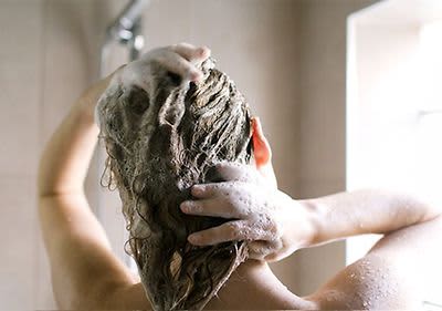 Osoba si myje vlasy ve sprše s pěnou na hlavě