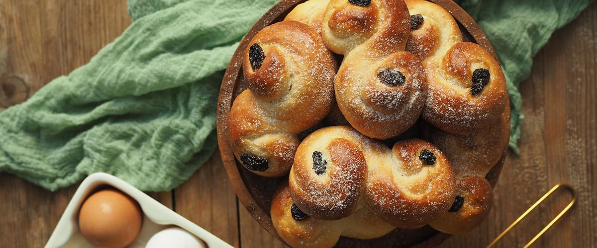 Pečivo s rozinkami na talíři, vedle krabička s vejci a zelená utěrka na dřevěném stole