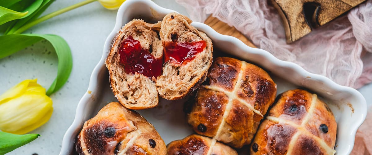 Pečivo s rozinkami a křížem, rozkrojené s marmeládou, žluté tulipány, dřevěný podnos, růžová látka