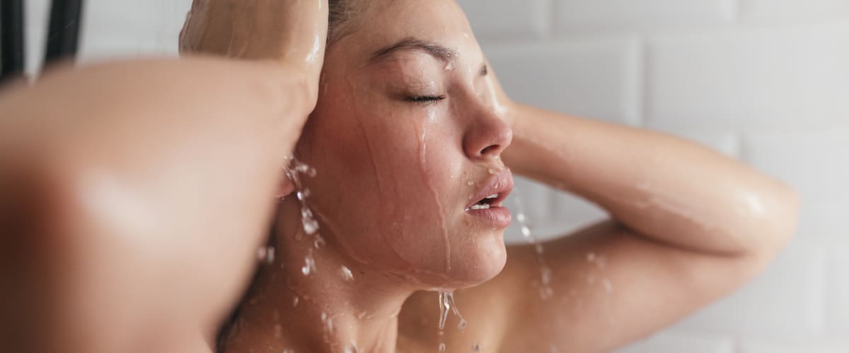 Osoba ve sprše s vodou stékající po těle a dlaždicemi na pozadí