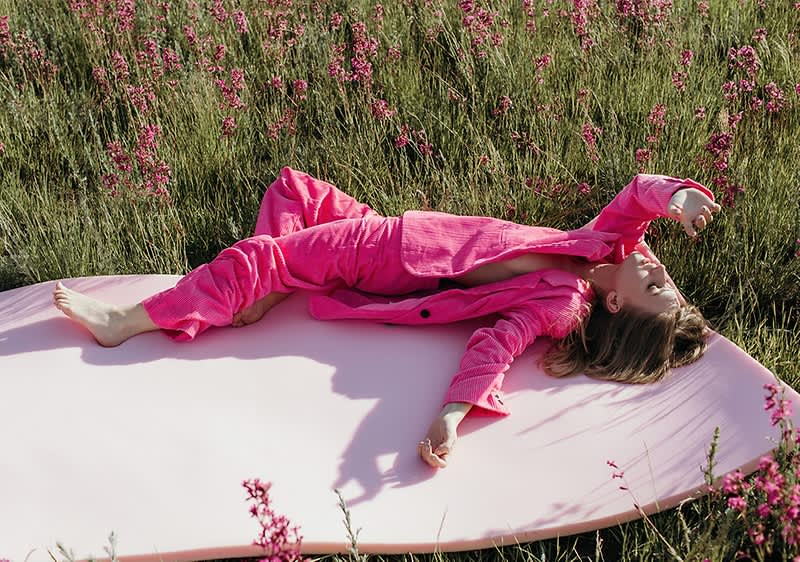 Žena v růžovém oblečení leží v poli s růžovými květinami.