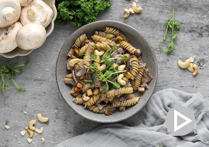 Těstoviny s houbami a kešu oříšky v misce na šedém stole, vedle čerstvé houby a petržel