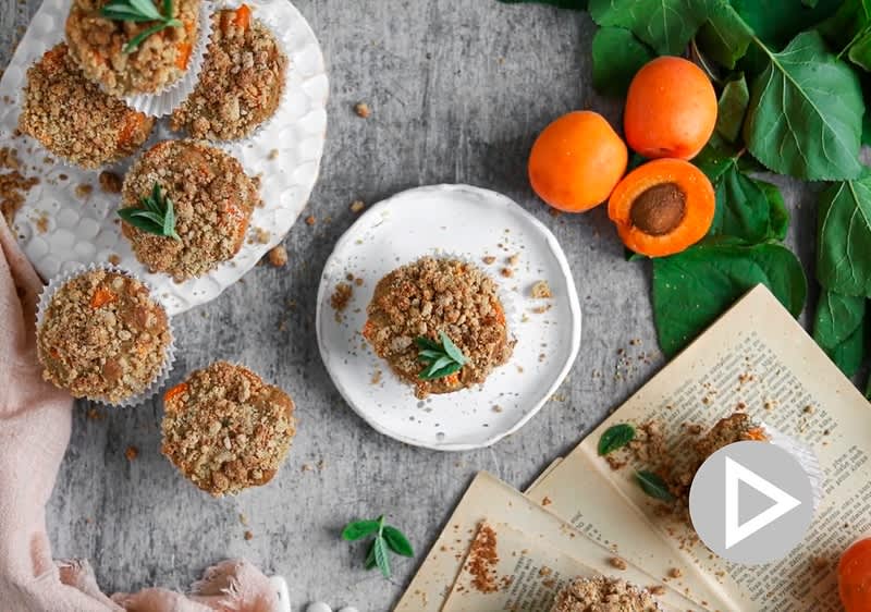 Muffiny s drobenkou na talířích, čerstvé meruňky, zelené listy a otevřená kniha na šedém povrchu