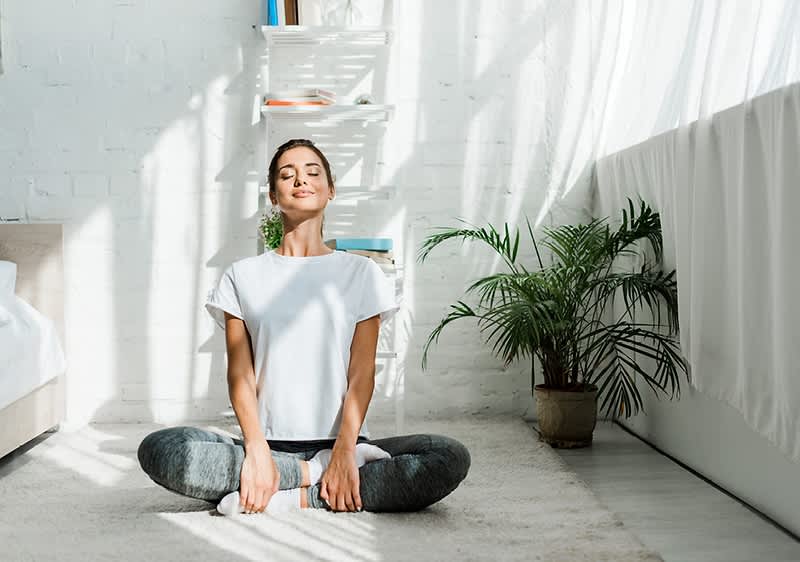 Žena sedící v meditaci v světlé místnosti s rostlinou a policí v pozadí