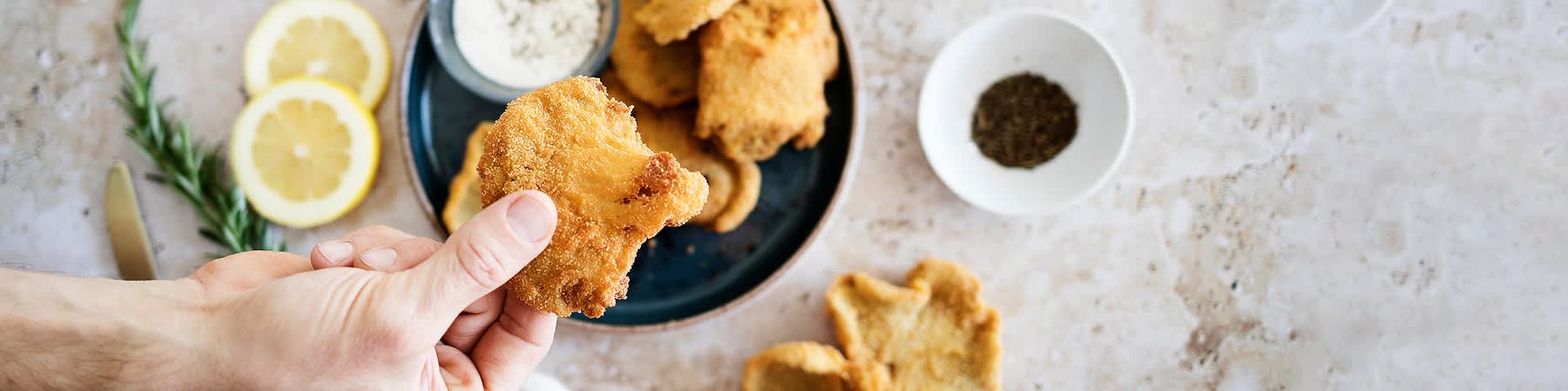 Smažené houby držené v ruce, talíř s dalšími houbami a dipem, plátky citronu na stole