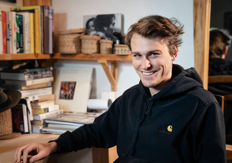 Muž v černé mikině sedící u stolu s knihami a košíky na poličce v pozadí