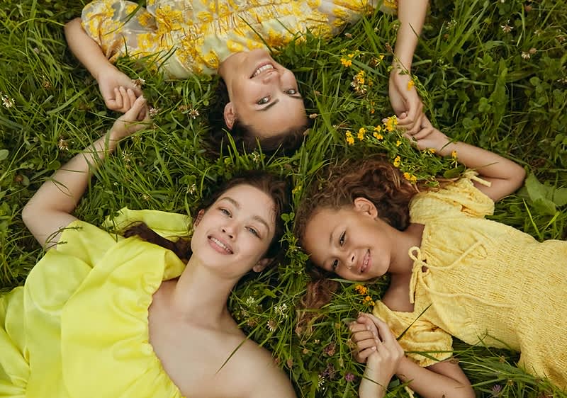 Tři osoby v žlutém oblečení leží na trávě, drží se za ruce, obklopeny květinami.