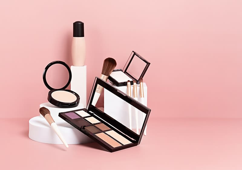 Kosmetické produkty na růžovém pozadí, včetně paletky očních stínů, pudru, štětců a lahvičky.