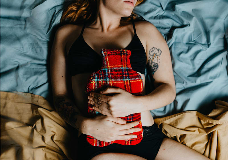 Žena ležící na posteli drží na břiše termoforovou láhev