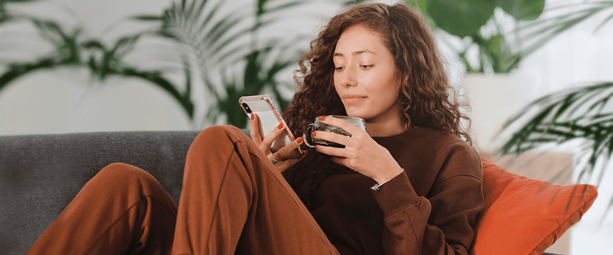 Žena drží telefon a šálek, sedí na pohovce