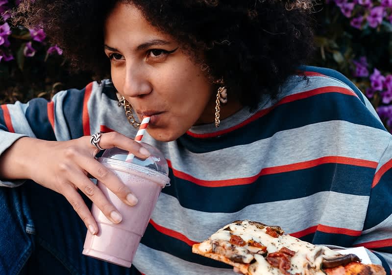 Osoba v pruhovaném svetru drží smoothie a kousek pizzy před květinami