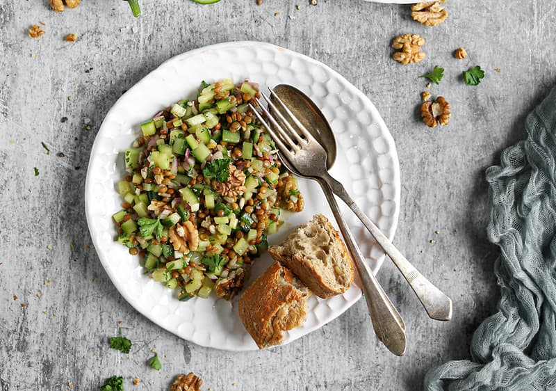Talíř s čerstvým salátem z okurky a ořechů, vedle kousek chleba a příbor na šedém pozadí