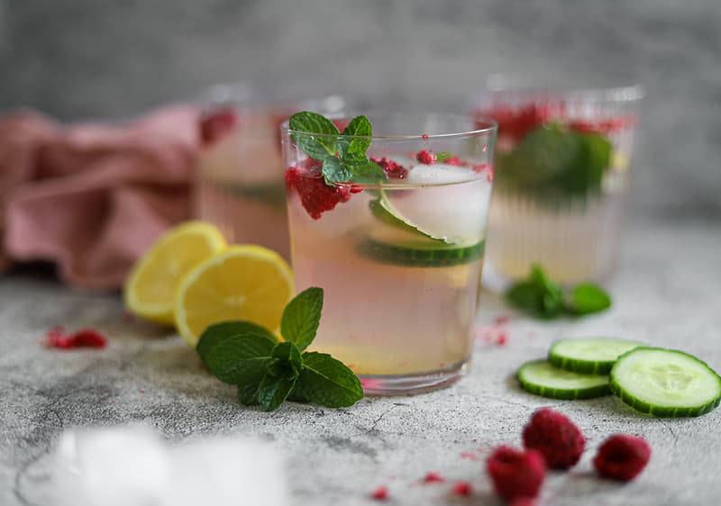 Sklenice s limonádou, plátky citronu, okurky, malinami a mátou na šedém pozadí