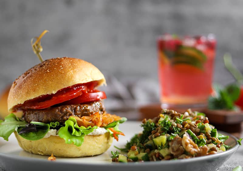 Burger se zeleninou na talíři, vedle salát, v pozadí sklenice s nápojem