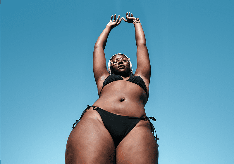 Žena v černých plavkách s rukama nad hlavou před modrou oblohou.