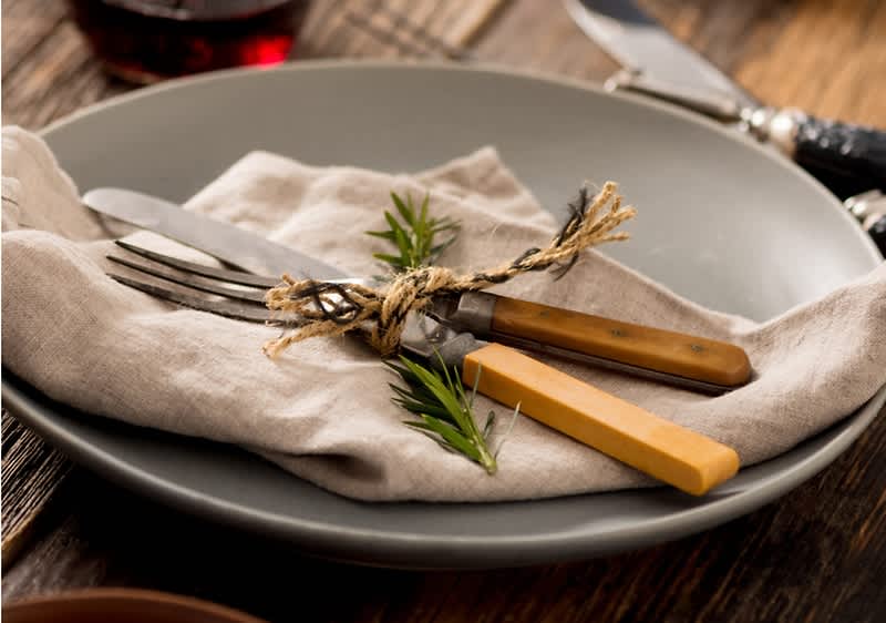 Talíř s příbory na látkovém ubrousku, ozdobený větvičkou rozmarýnu na dřevěném stole.