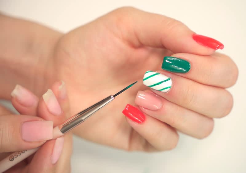 Ruka s barevnými nalakovanými nehty, jeden nehet s pruhy, štětec na nehty v druhé ruce