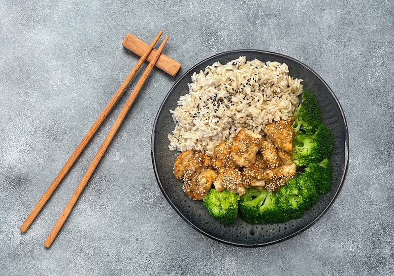 Talíř s rýží, kuřecím masem a brokolicí, vedle dřevěné hůlky na šedém pozadí