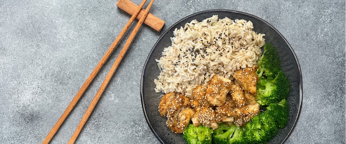 Talíř s rýží, kuřecím masem a brokolicí, vedle dřevěné hůlky na šedém pozadí