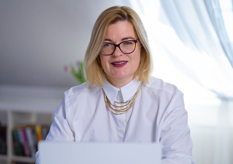 Žena v bílé košili sedící u stolu s notebookem, v pozadí knihovna a okno s květinou.