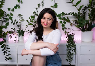 Žena sedící před bílým nábytkem, obklopená zelenými rostlinami na pozadí.