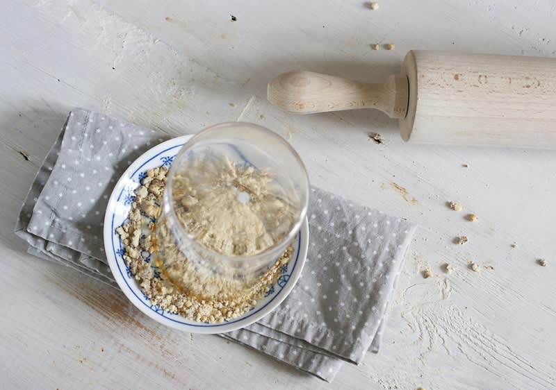 Sklenice na talíři s ovesnými vločkami, vedle váleček na těsto na světlém stole.