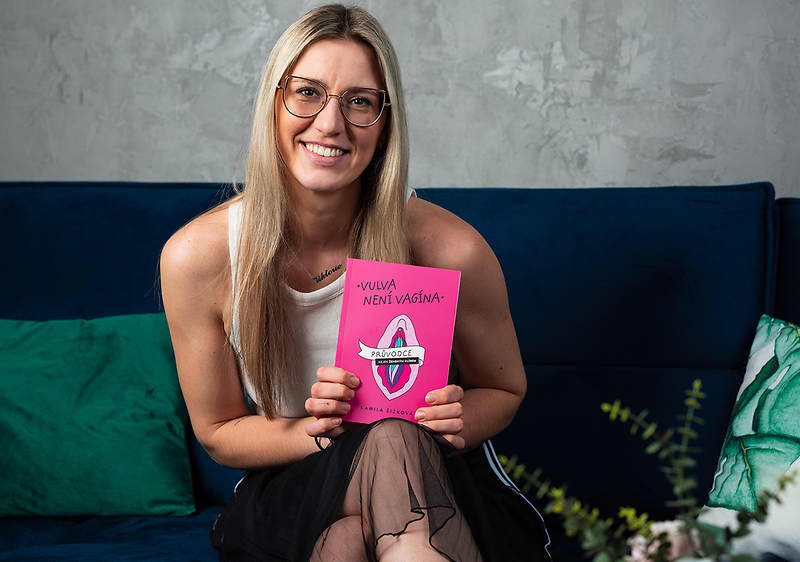 Žena sedící na pohovce drží růžovou knihu s nápisem Vulva není vagína