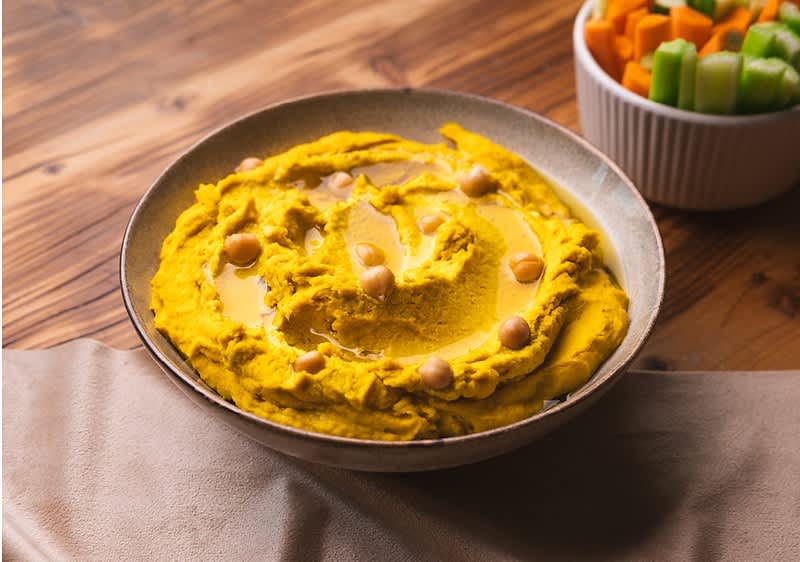 Talíř hummusu s cizrnou na dřevěném stole, vedle miska s nakrájenou zeleninou