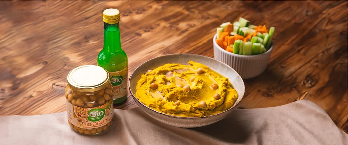 Talíř hummusu s cizrnou na dřevěném stole, vedle miska s nakrájenou zeleninou