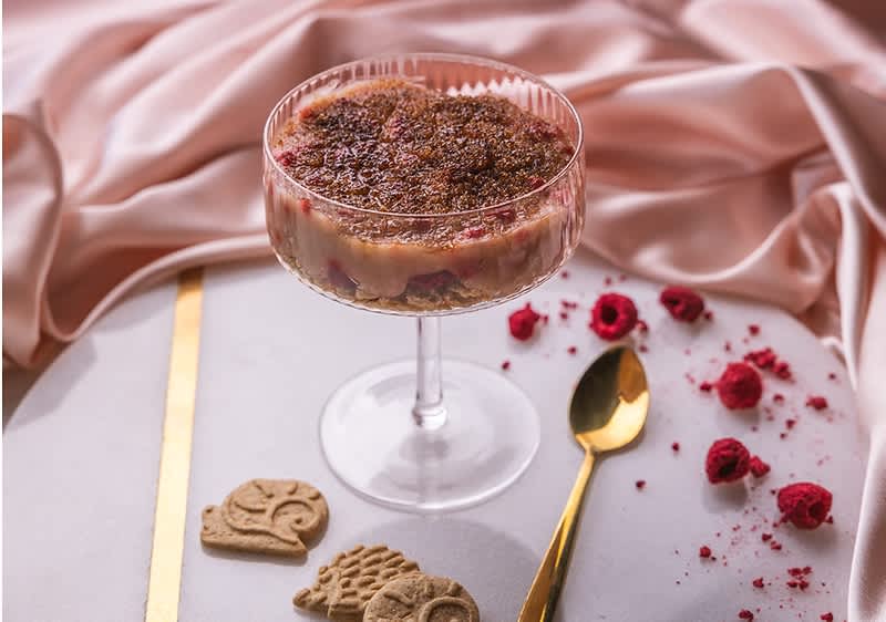 Dezert v sklenici s malinami, zlatá lžička, sušenky a růžová látka na pozadí