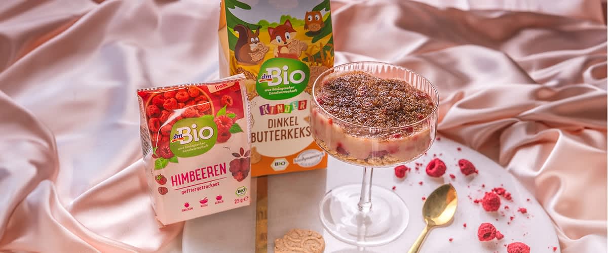 Dezert v sklenici s malinami, zlatá lžička, sušenky a růžová látka na pozadí