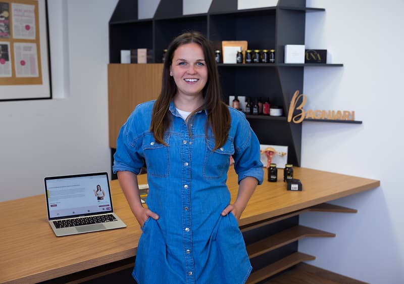 Žena v modré košili stojí u stolu s notebookem a kosmetickými produkty na poličkách