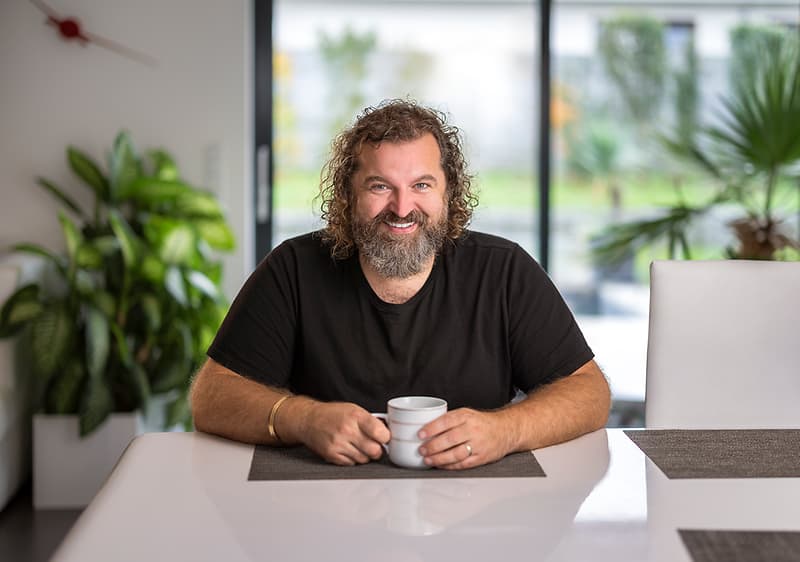 Muž sedící u stolu s hrnkem, v pozadí rostliny a velké okno