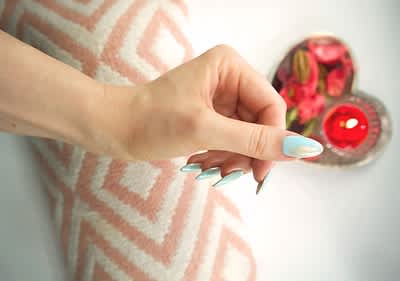 Ruka s modrými nehty na vzorovaném polštáři vedle srdcového talíře s květinami a svíčkou