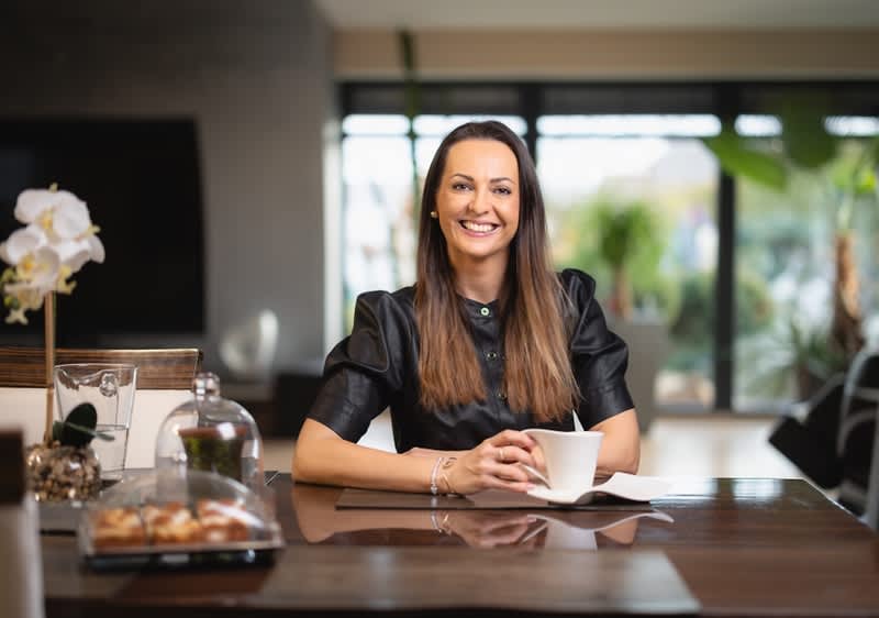 Žena sedící u stolu s šálkem, rozmazaný obličej, v pozadí rostliny a moderní interiér.