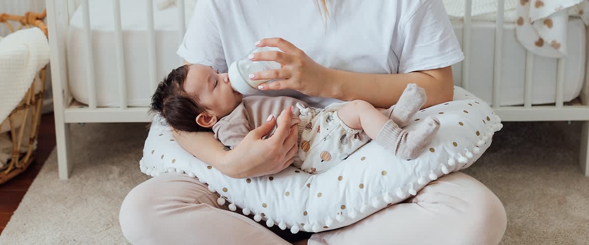 Žena krmí kojence lahví, dítě leží na polštáři v náručí ženy.