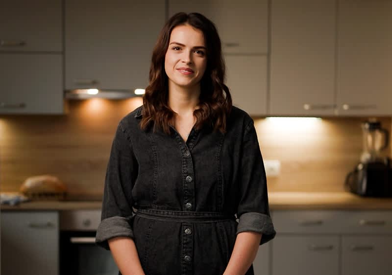 Žena stojící v kuchyni s rozmazaným obličejem, za ní jsou skříňky a kuchyňské spotřebiče.