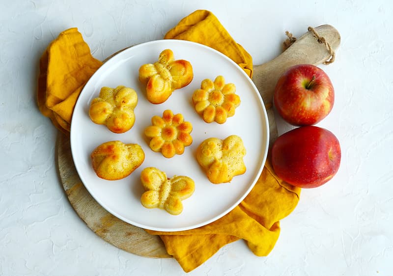 Pečené muffiny ve tvaru květin a motýlů na talíři vedle dvou červených jablek