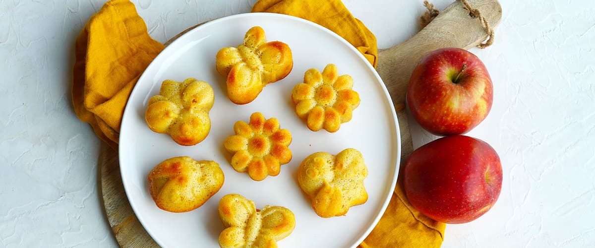 Pečené muffiny ve tvaru květin a motýlů na talíři vedle dvou červených jablek